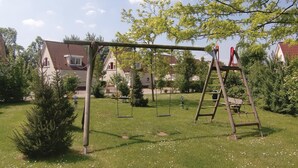 Un terrain de jeux avec des balançoires et une échelle, entouré d’herbe et d’arbres, avec des maisons en arrière-plan.