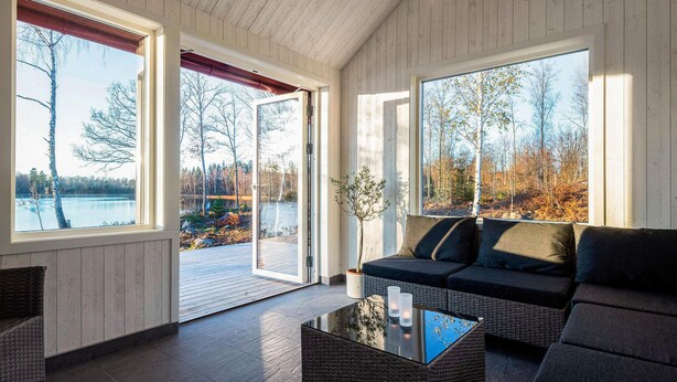 En modern inredning med en soffa, ett soffbord i glas och stora fönster med utsikt över en sjö och träd.