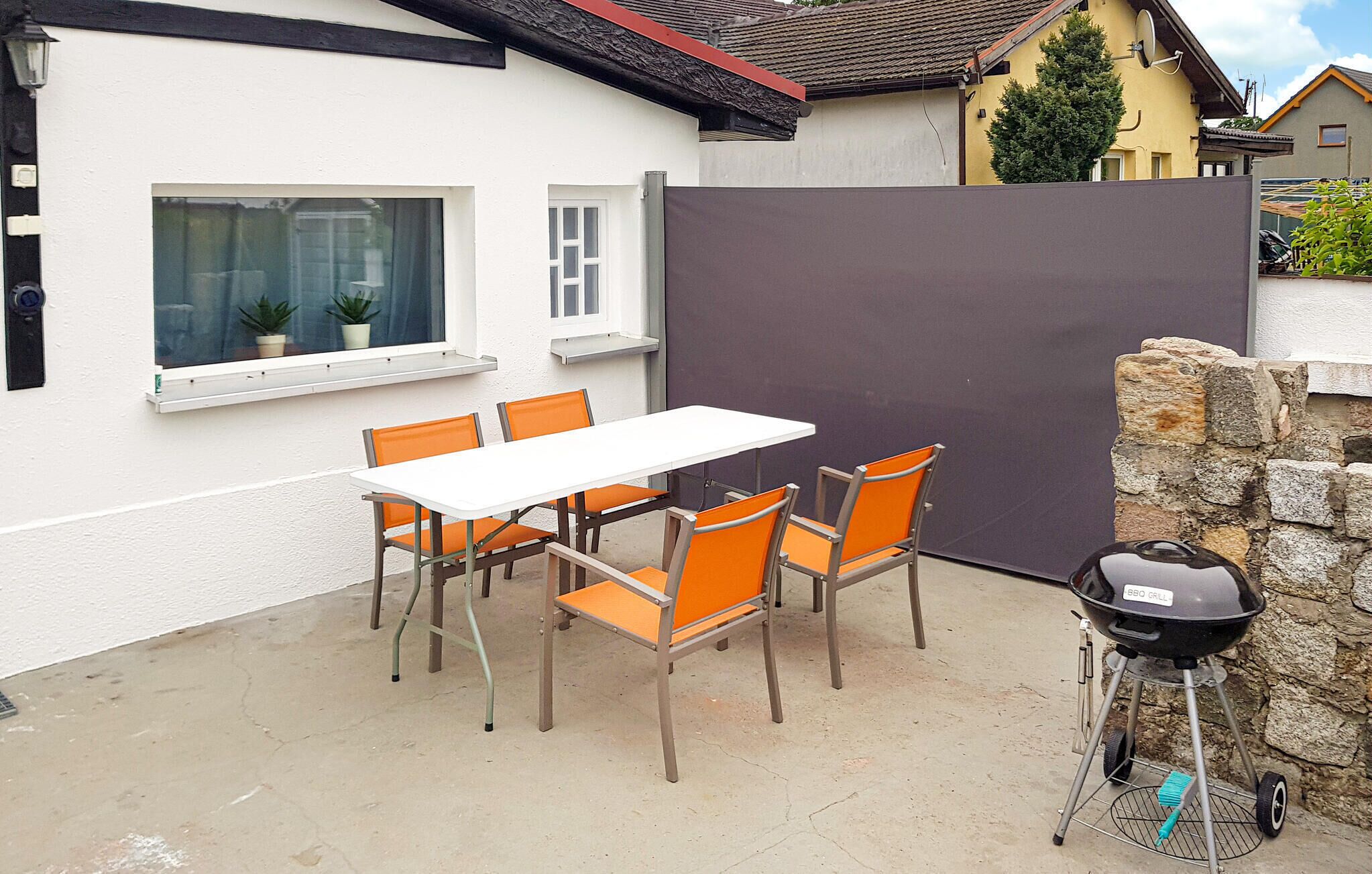 A patio with a white table and orange chairs, a barbecue grill, and a stone wall.