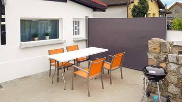 A patio with a white table and orange chairs, a barbecue grill, and a stone wall.