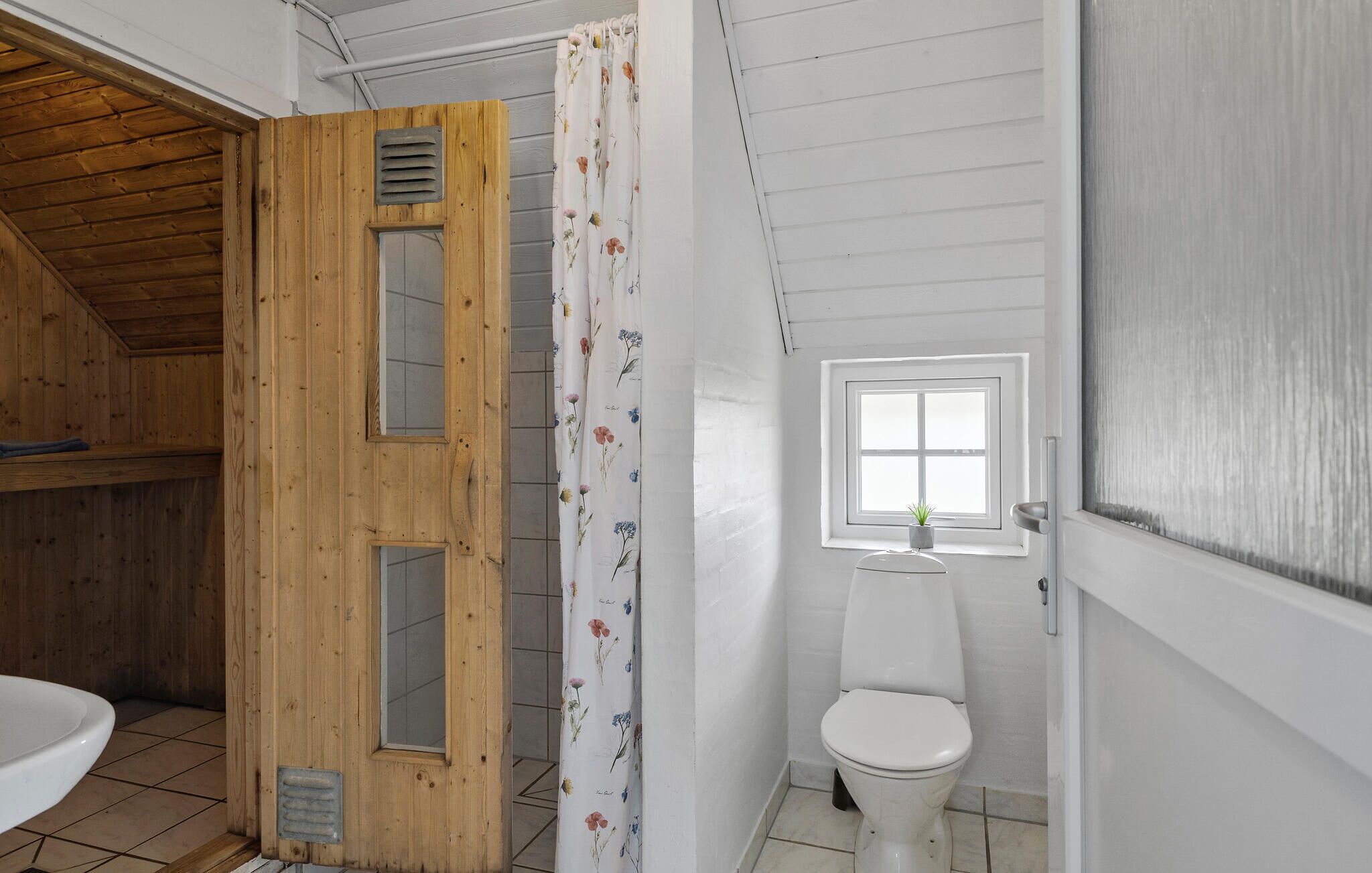 Une salle de bain avec une porte en bois, un rideau de douche orné de motifs floraux, des toilettes blanches et une petite fenêtre avec une plante.