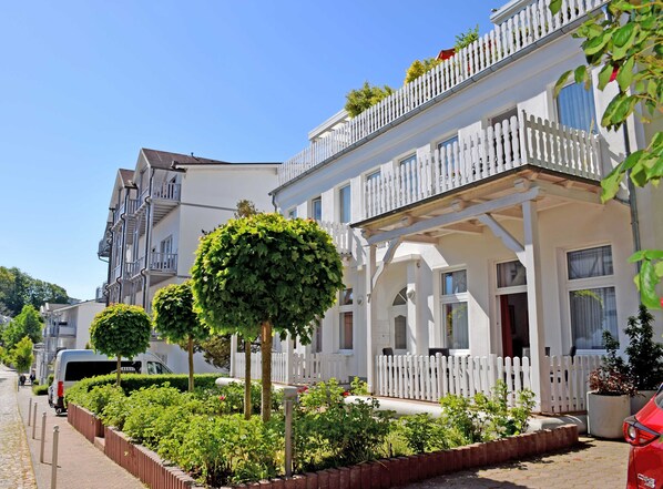 A row of white two-story buildings with balconies, a paved street, and neatly trimmed hedges.