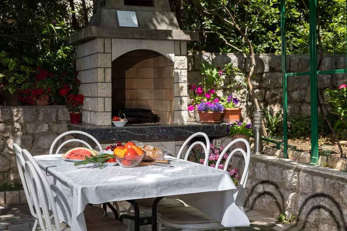An outdoor dining area with a stone-built oven, white metal chairs, and a table set with fruits and vegetables.