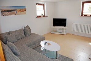 Un salon avec un canapé gris, une table basse blanche, un téléviseur à écran plat posé sur un meuble blanc et un tableau sur le mur représentant un paysage de plage.