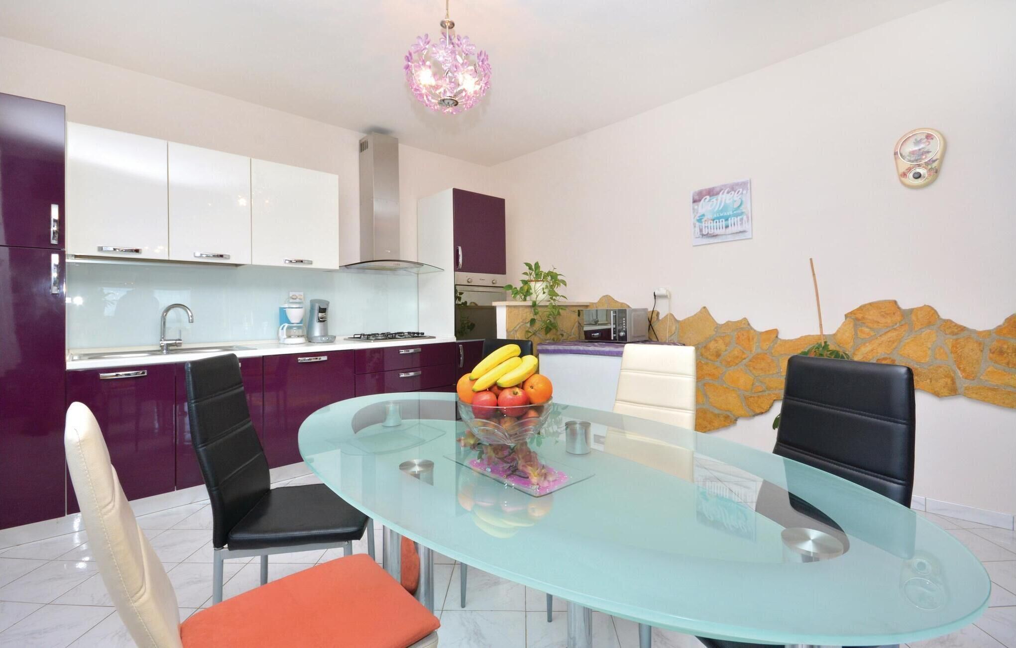 Een moderne keuken met witte kasten, een gasfornuis en een koffiezetapparaat. Er is een zithoek met een glazen tafel en stoelen, en een fruitschaal op de tafel.