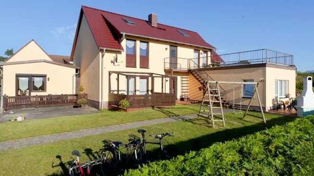 A two-story house with a red roof, a visible staircase, and a small garden area with a bicycle.