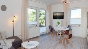 A modern living room with a sofa, a dining table with chairs, a TV mounted on the wall, and large windows.