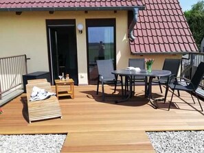 Una terraza de madera con una mesa, sillas y una tumbona, y una puerta que da a un espacio interior.