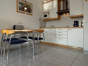 A modern kitchen with white cabinets, a stainless steel range hood, a microwave, and a coffee maker. There is a wooden table with chairs and a hanging light fixture.