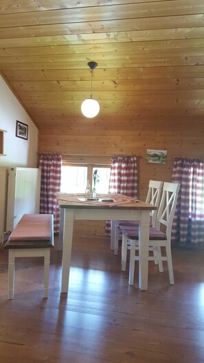 A wooden interior with a dining table, chairs, a radiator, and checkered curtains.