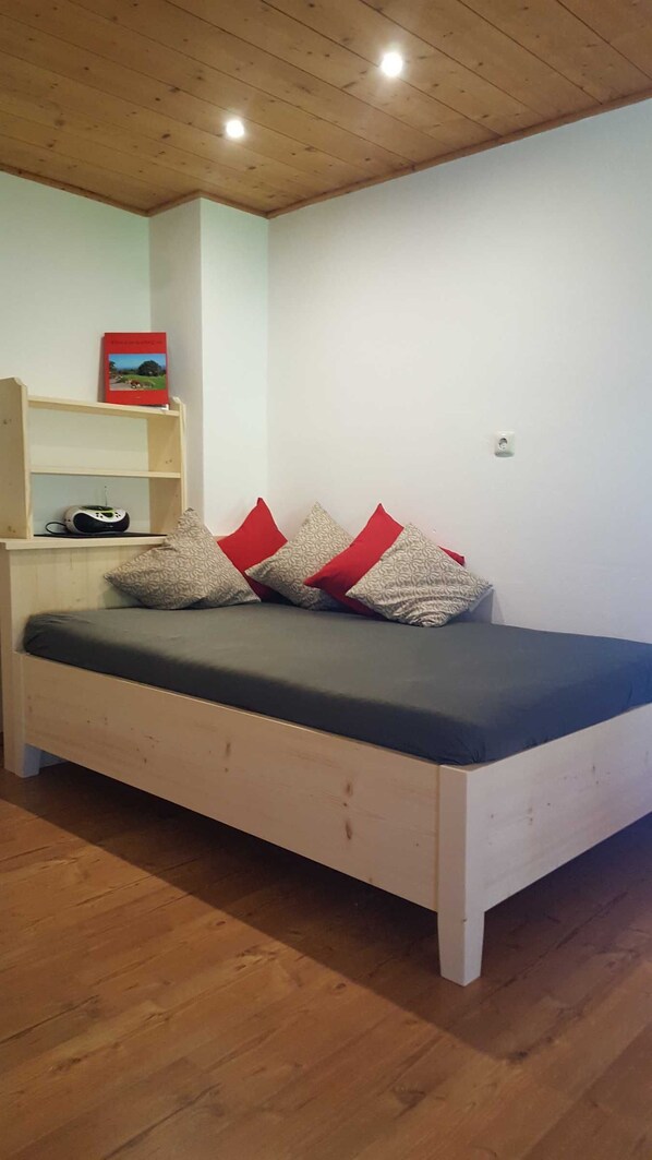 Un lit simple avec un matelas gris, une tête de lit en bois et des oreillers rouges et gris. Une étagère avec un livre et un appareil se trouve à gauche.