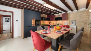Un comedor moderno con una mesa de madera dispuesta para cuatro personas, rodeada de sillas rojas y grises. Se ve una cocina con armarios oscuros y una pared de piedra al fondo.