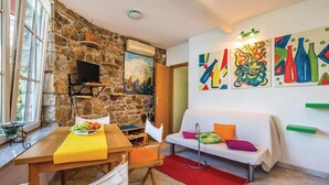 Un salon chaleureux avec un mur en pierre, une table en bois sur laquelle sont disposés des fruits, un canapé blanc et des œuvres d’art aux couleurs vives.