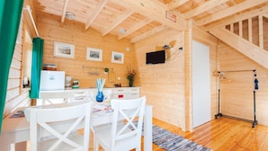 L’intérieur d’un chalet en bois accueillant, avec un coin repas, des chaises blanches, une petite table, un réfrigérateur, un téléviseur et une porte donnant sur une autre pi èce.