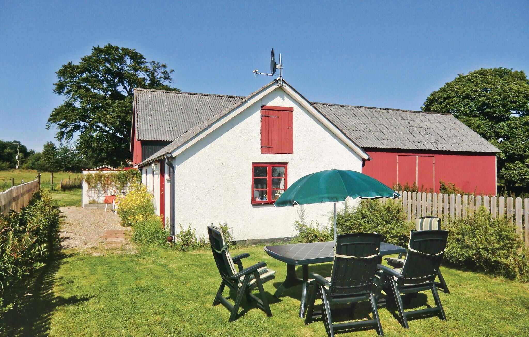 A white house with a red door and window shutters, a red barn in the background, and a table with chairs in the front yard.