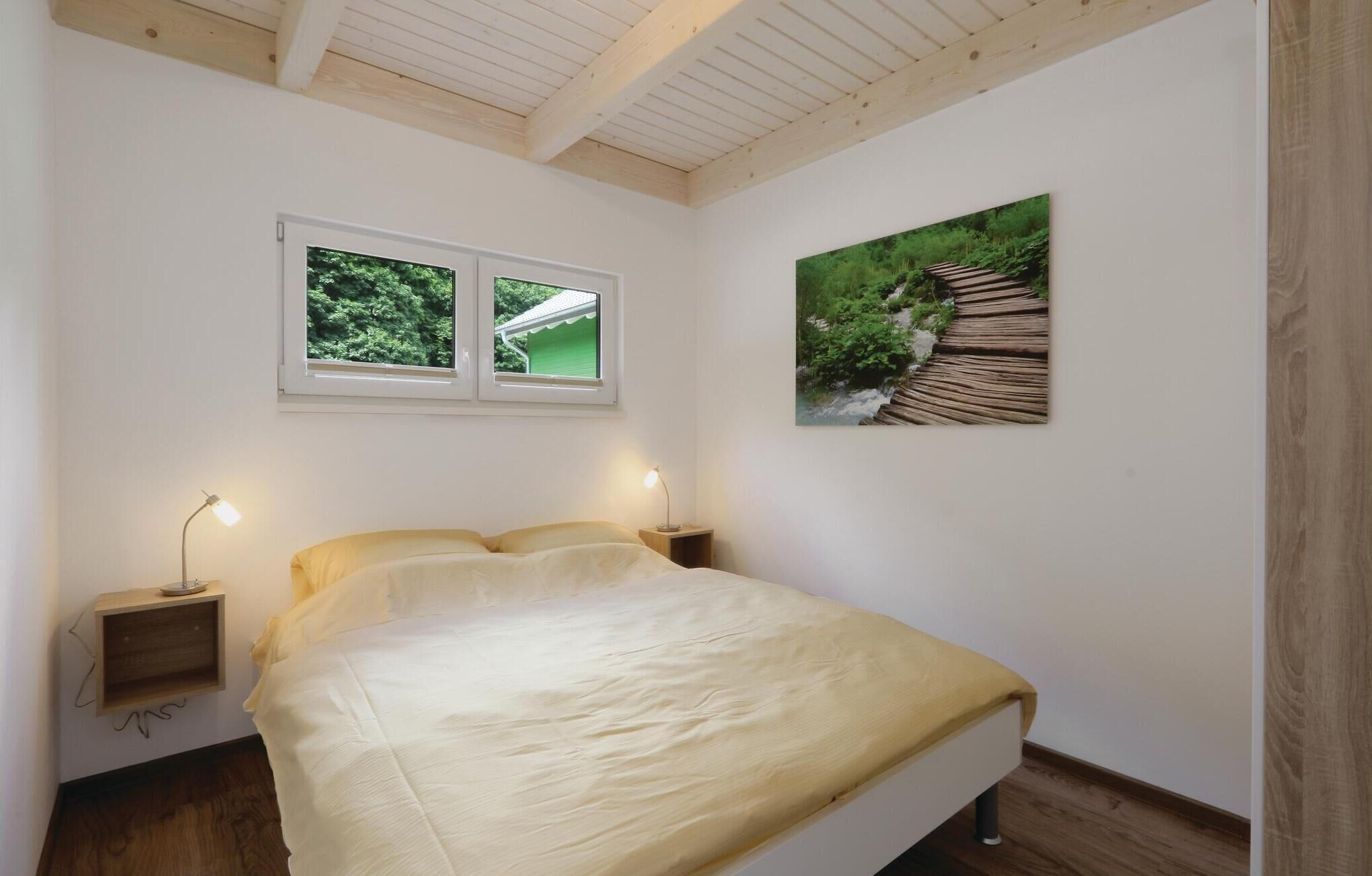 Une chambre à coucher avec un lit, deux tables de chevet, une fenêtre donnant sur un paysage verdoyant et un tableau représentant un pont en bois.
