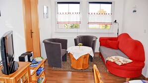 Ett vardagsrum med en tv, två fåtöljer, en röd soffa, ett litet bord med en vas och fönster med gardiner.