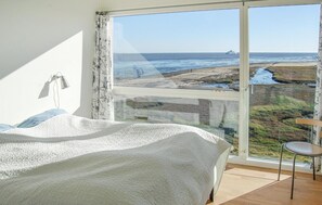 Ein Schlafzimmer mit großem Fenster mit Blick auf Strand und Meer, ein Bett und ein kleiner Tisch.