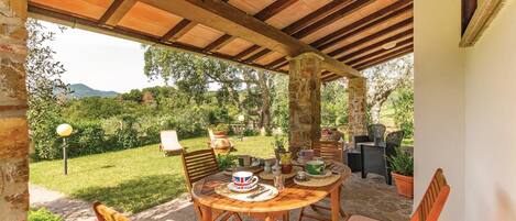 An outdoor patio with a wooden table and chairs, surrounded by stone pillars and a view of a lush garden.