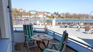 Un balcon avec deux chaises à rayures vertes et une petite table ronde, donnant sur une zone riveraine avec des bateaux et des bâtiments.