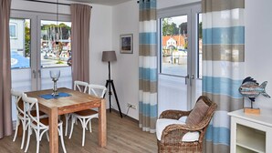 Une pièce comprenant une table à manger en bois, des chaises blanches, un fauteuil en osier, une lampe et une vue sur un port de plaisance.