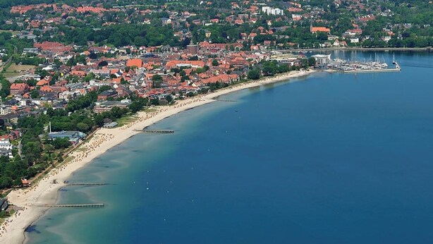 Vue aérienne d’une ville côtière avec une plage, un port de plaisance et des bâtiments aux toits rouges.