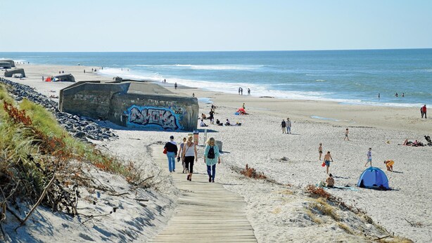 Een strand met een houten looppad dat leidt naar een grote rots met graffiti, mensen die lopen en tenten.
