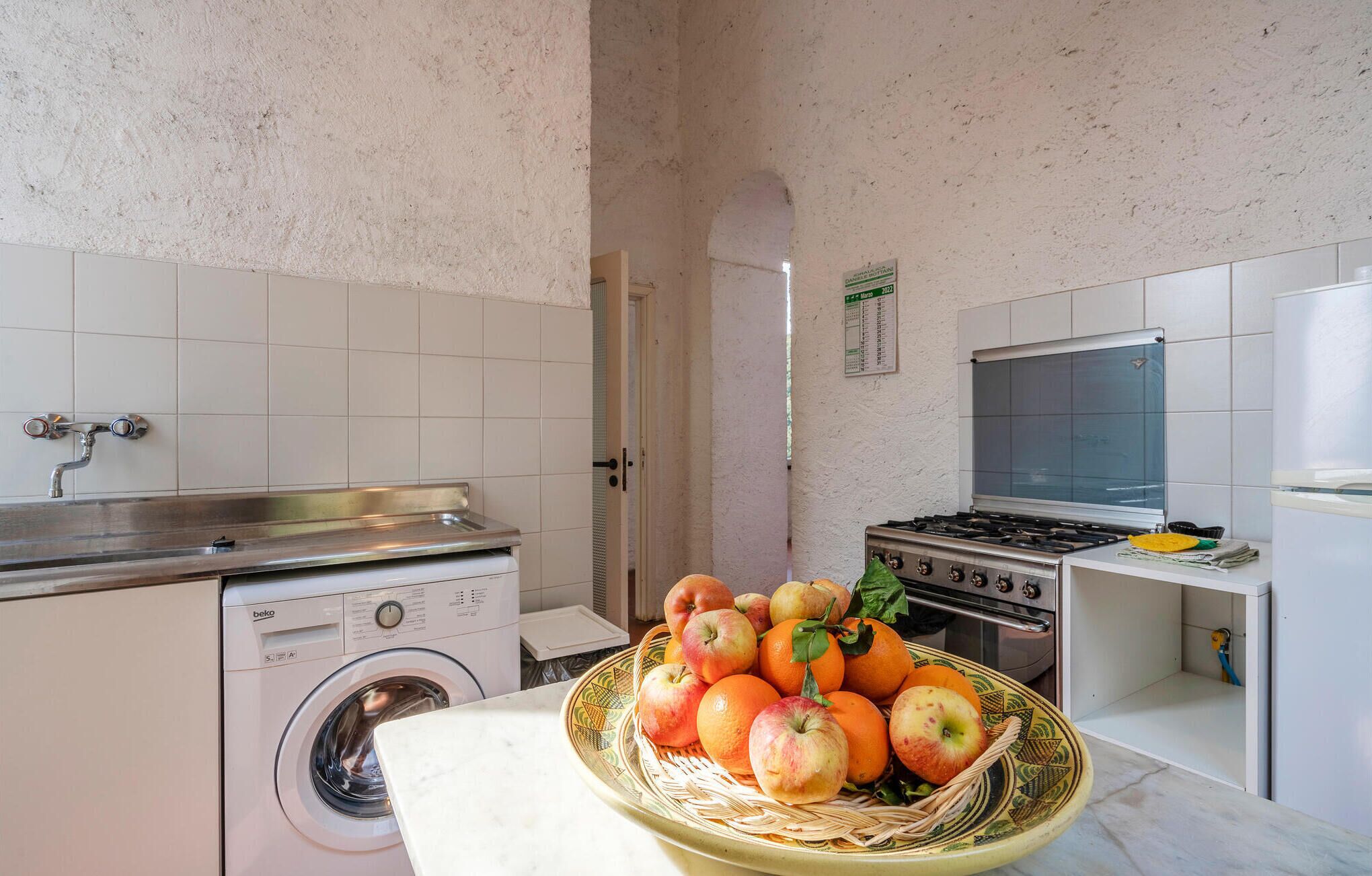 A kitchen with a washing machine, a stove, and a sink. There is a basket of fruit on the counter.