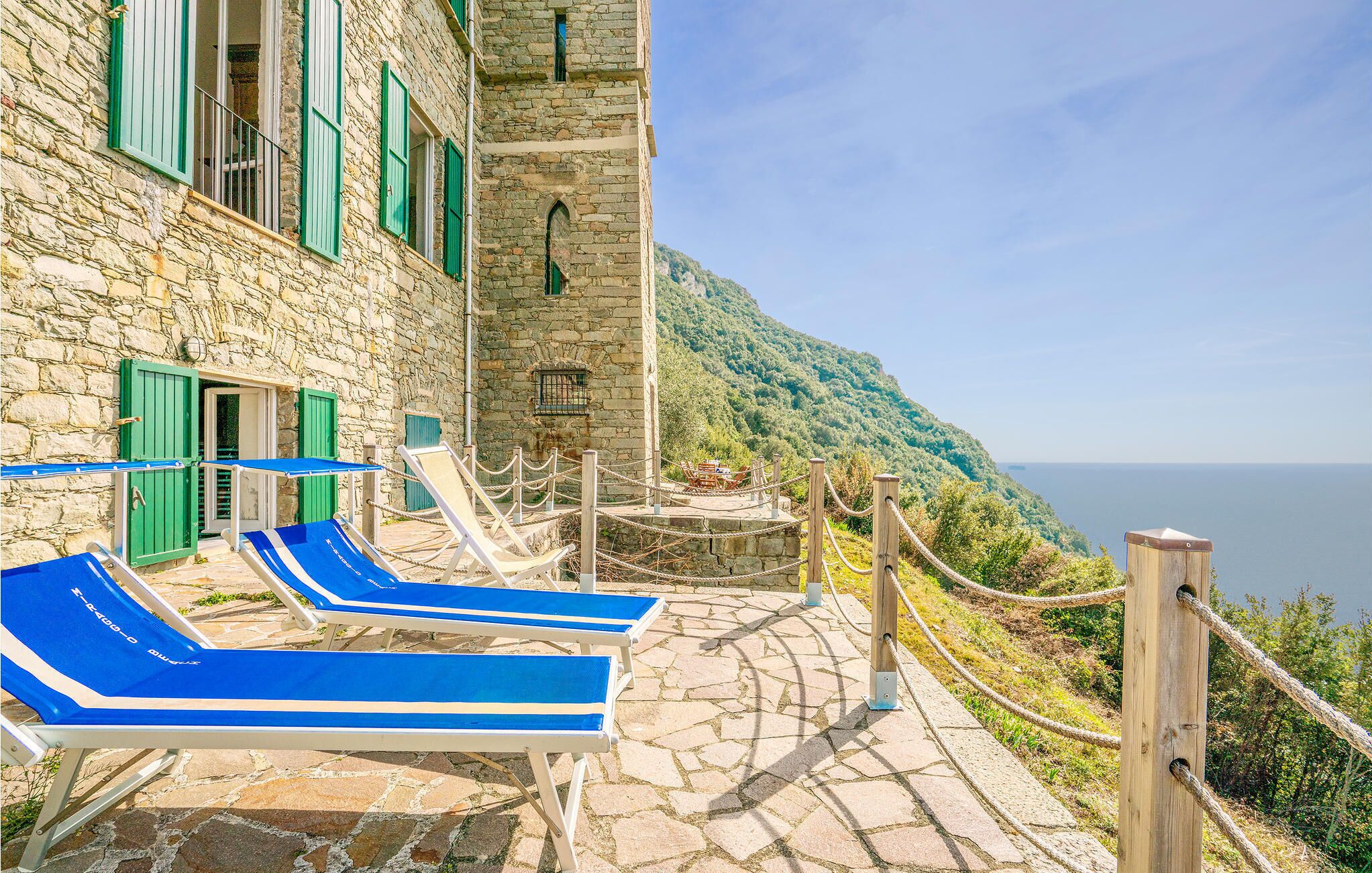 Un edificio in pietra con persiane verdi, una terrazza in pietra con poltrone relax e vista sul mare.