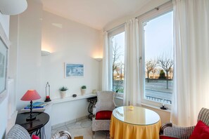 Una habitación con una mesa redonda de vidrio, una silla roja, una lámpara, una estantería, un ventanal con cortinas y un cuadro en la pared.