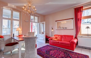 Una sala de estar con un sofá rojo, una mesa de comedor con sillas, una araña y un ventanal con cortinas.