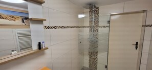 Un bagno con piastrelle bianche, una doccia con porta in vetro e uno scaffale con articoli da toilette.