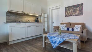 Eine moderne Küche mit weißen Schränken, einer Holzarbeitsplatte und einer Kaffeemaschine. Ein Esstisch mit blauem Tischläufer und einer Weinflasche steht bereit. Ein beiges Sofa mit Zierkissen lehnt an der Wand.