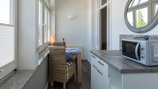 Eine kleine Küche mit einer Mikrowelle, einem Korbstuhl, einem Tisch und einem Fenster mit Jalousien.