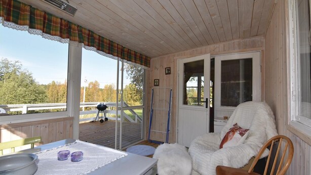A cozy room with a wooden ceiling, a plaid curtain, a window with a view of trees, a chair, a table with a lace tablecloth, and a grill outside. - Amazing home in årjäng with WiFi (Årjäng)