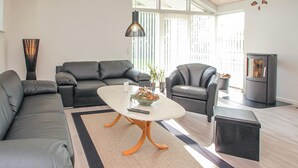Een moderne woonkamer met een modulaire bank, een salontafel en twee fauteuils. Er is een open haard en een groot raam met zonwering.