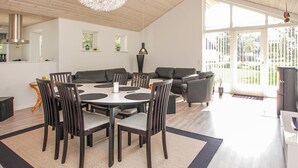 Een ruime woonkamer met een ronde eettafel omringd door stoelen, een bank en een groot raam met uitzicht op de omgeving.