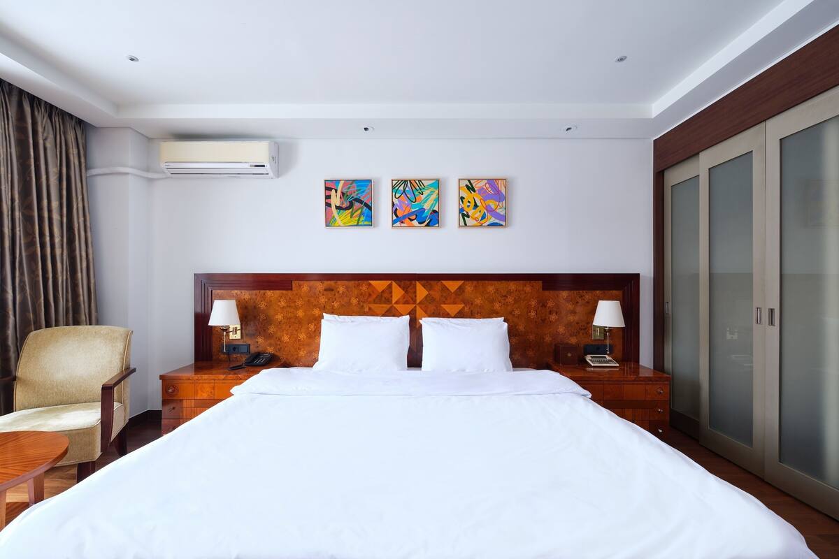 A hotel room with a large bed, a wooden headboard, a desk with a lamp, a chair, and a wardrobe.