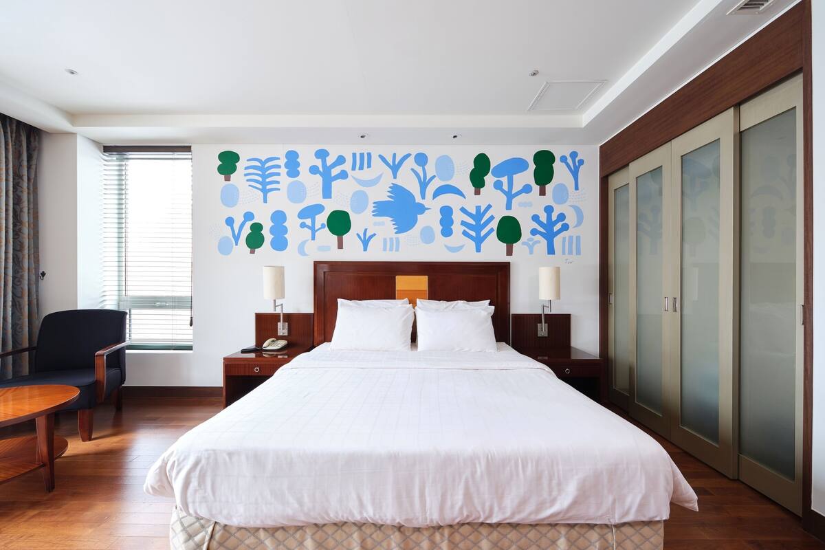 A hotel room with a large bed, two bedside tables with lamps, a chair, a wooden desk, and a wardrobe with frosted glass doors.