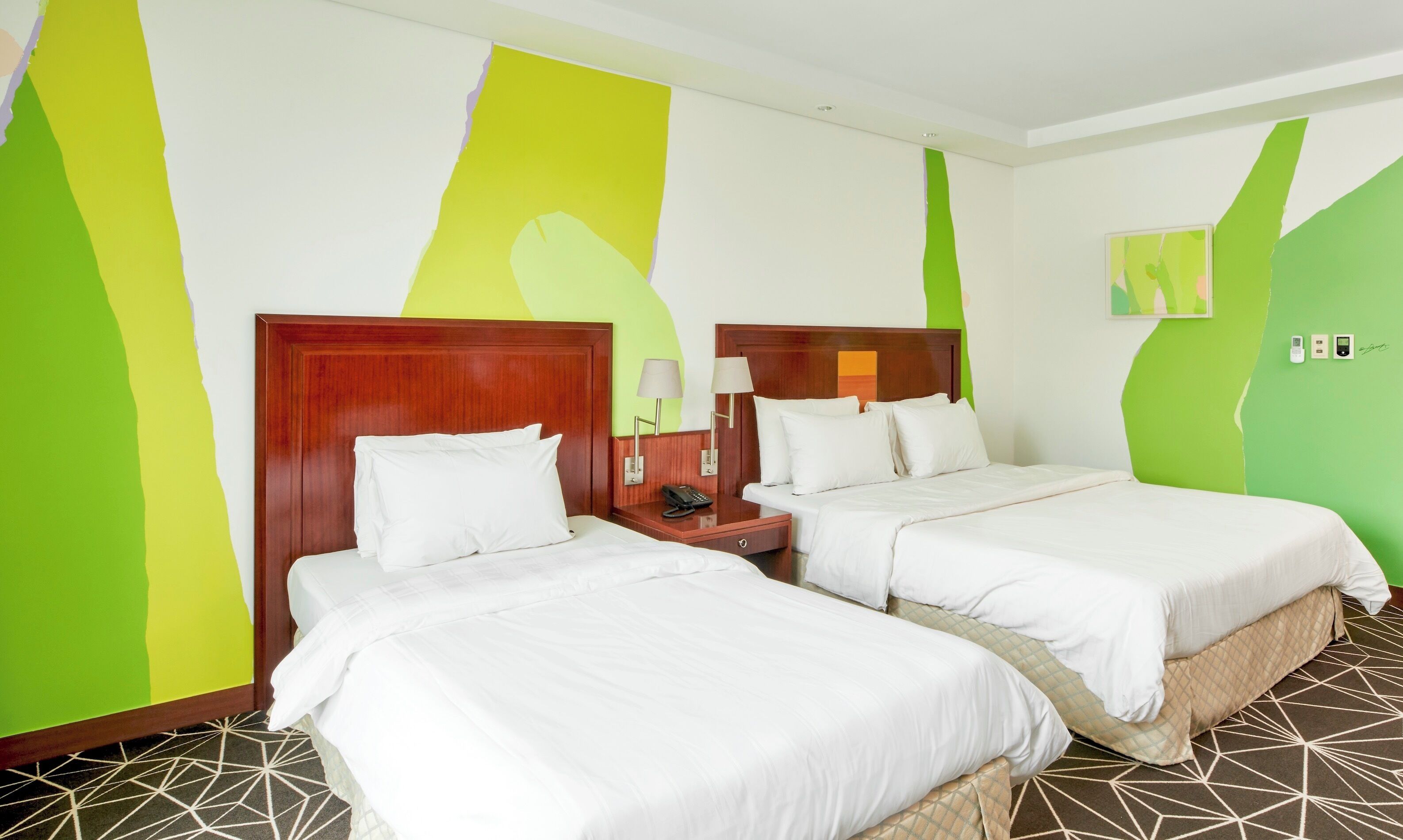 Habitación de hotel con dos camas, cabecero de madera, mesita de noche con lámpara y suelo con estampado geométrico.