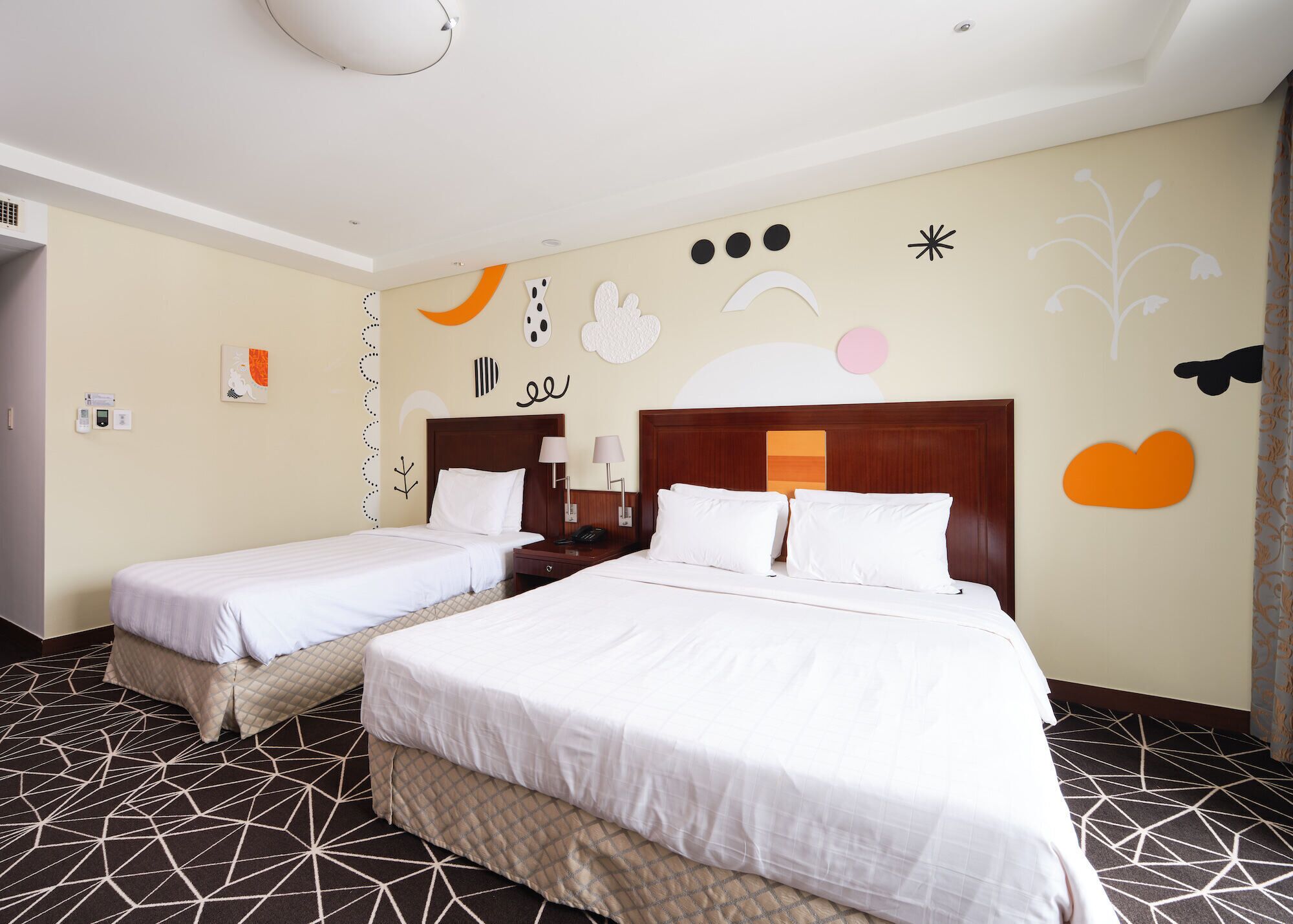 A hotel room with two beds, a wooden headboard, a nightstand, a lamp, and a wall with abstract art.