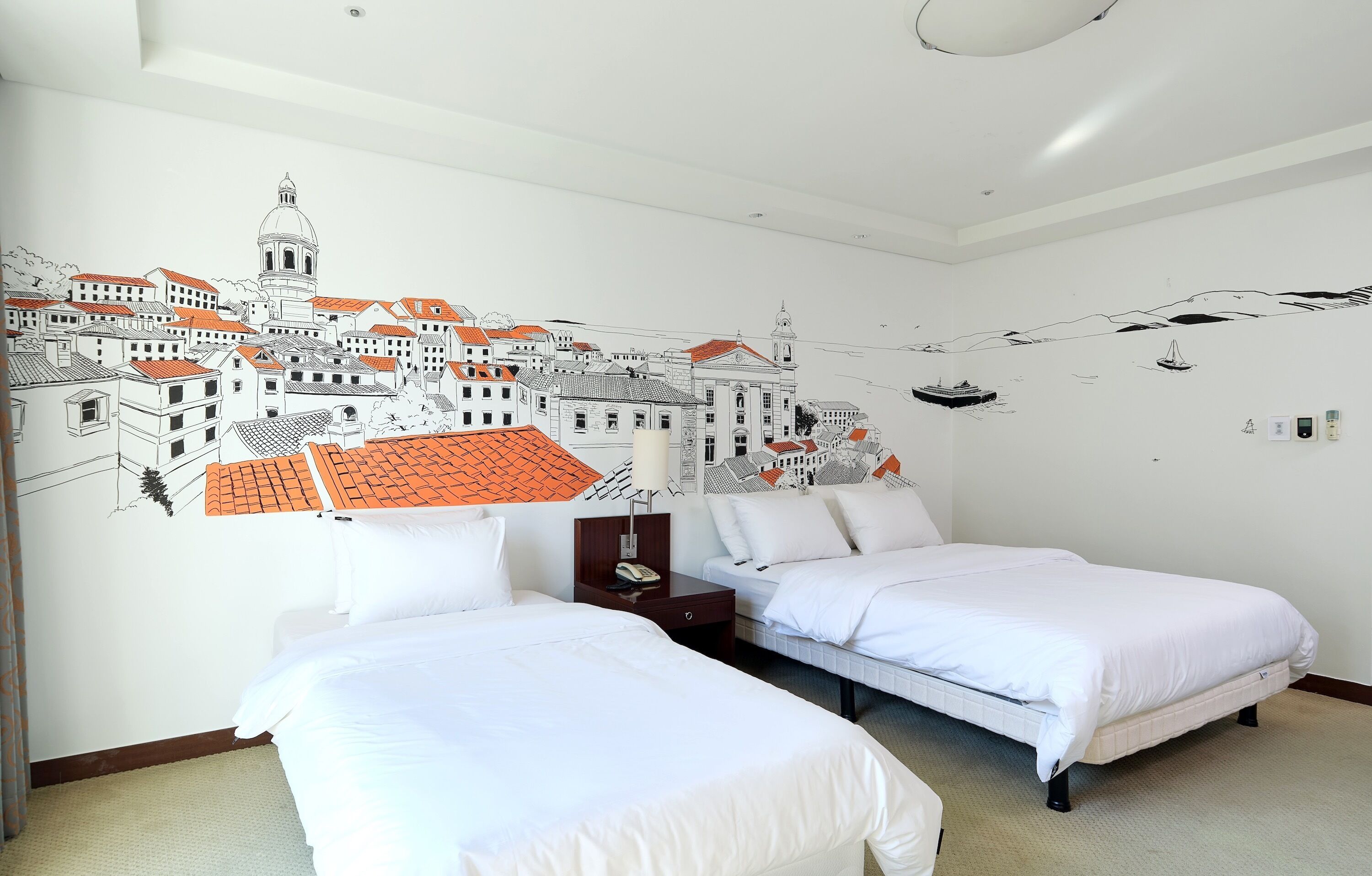 A hotel room with two beds, a wall mural of a town, and a small table with a lamp.