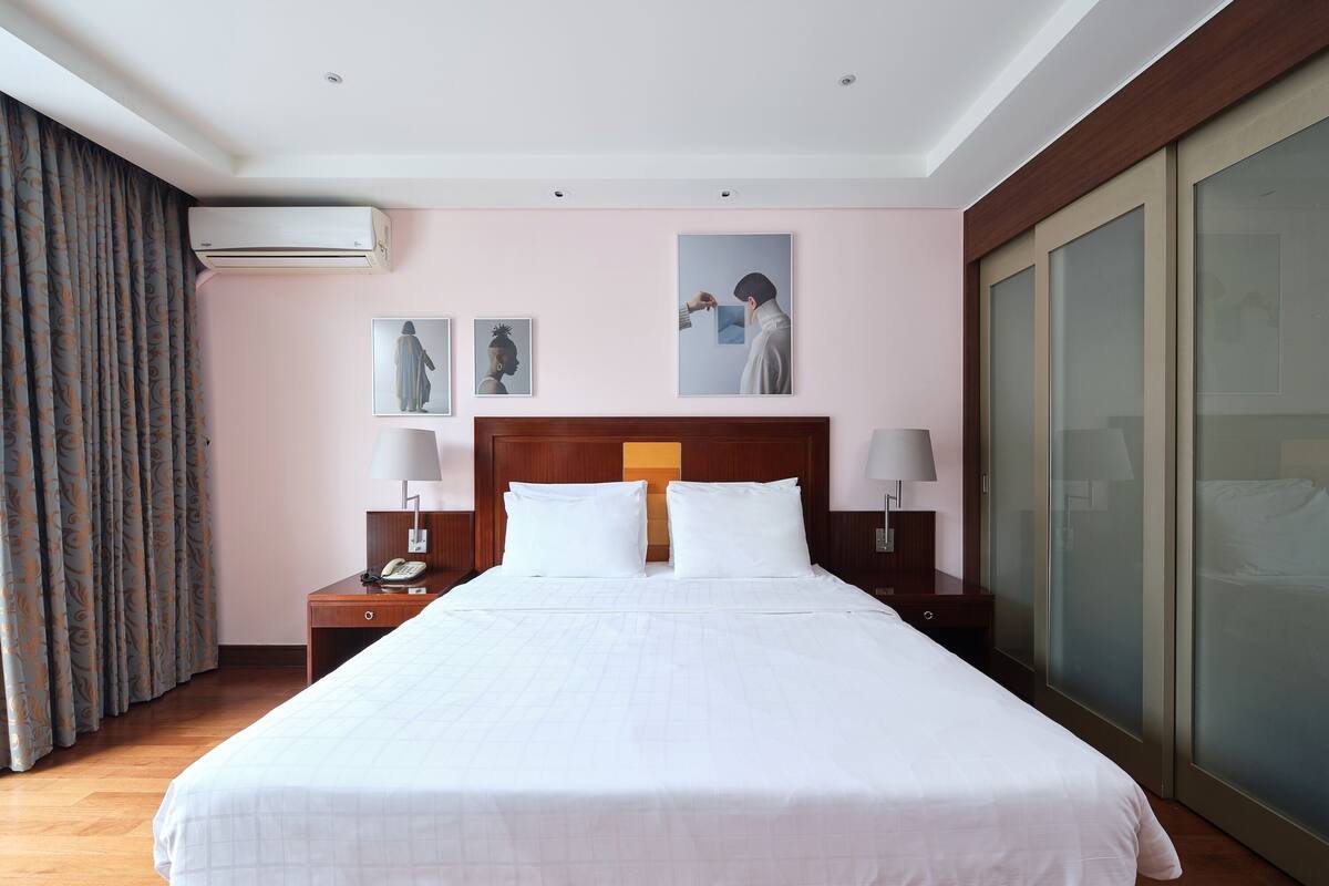 A hotel room with a large bed, two bedside tables with lamps, an air conditioning unit, and a sliding glass door closet.