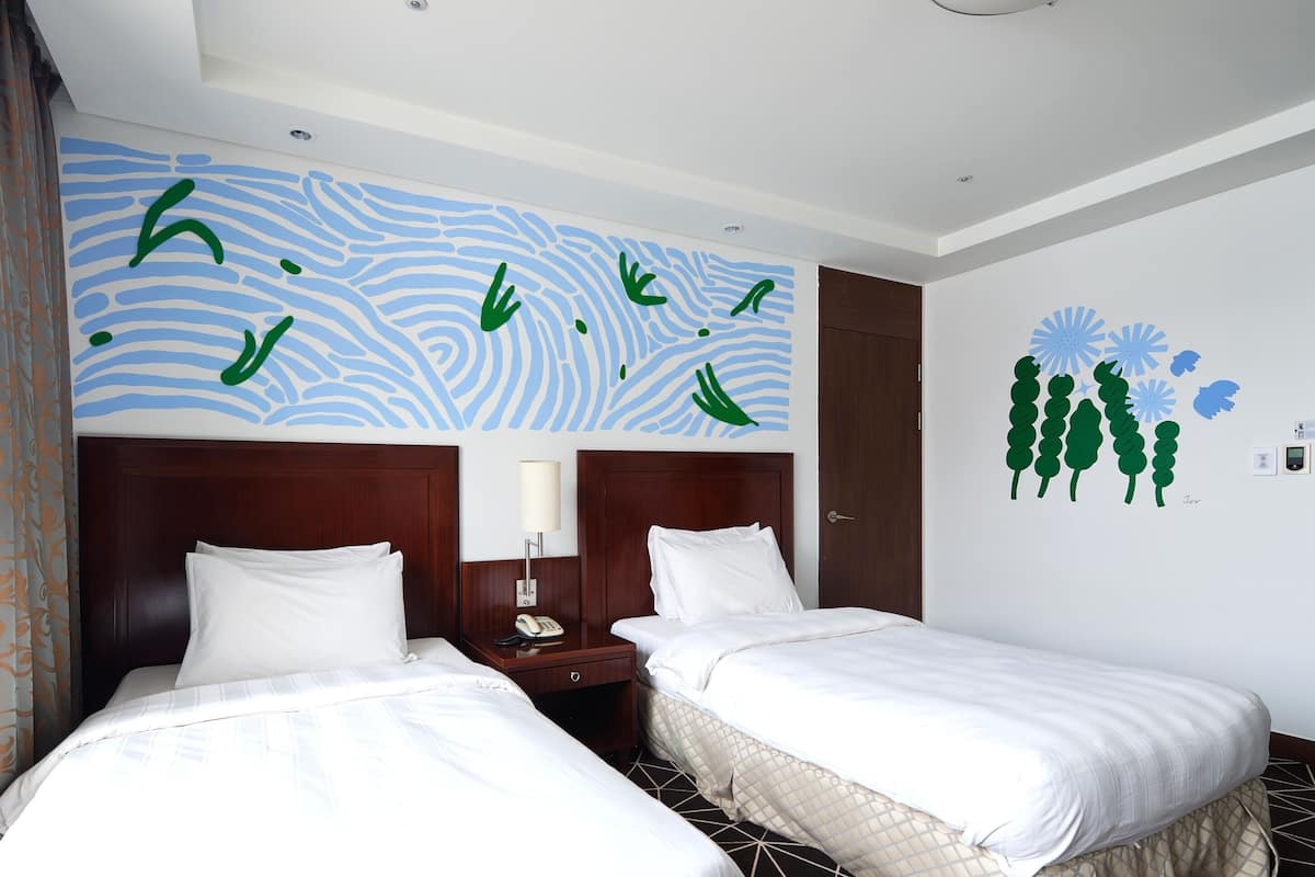 A hotel room with two beds, a nightstand with a lamp, a wall with abstract art, and a door.