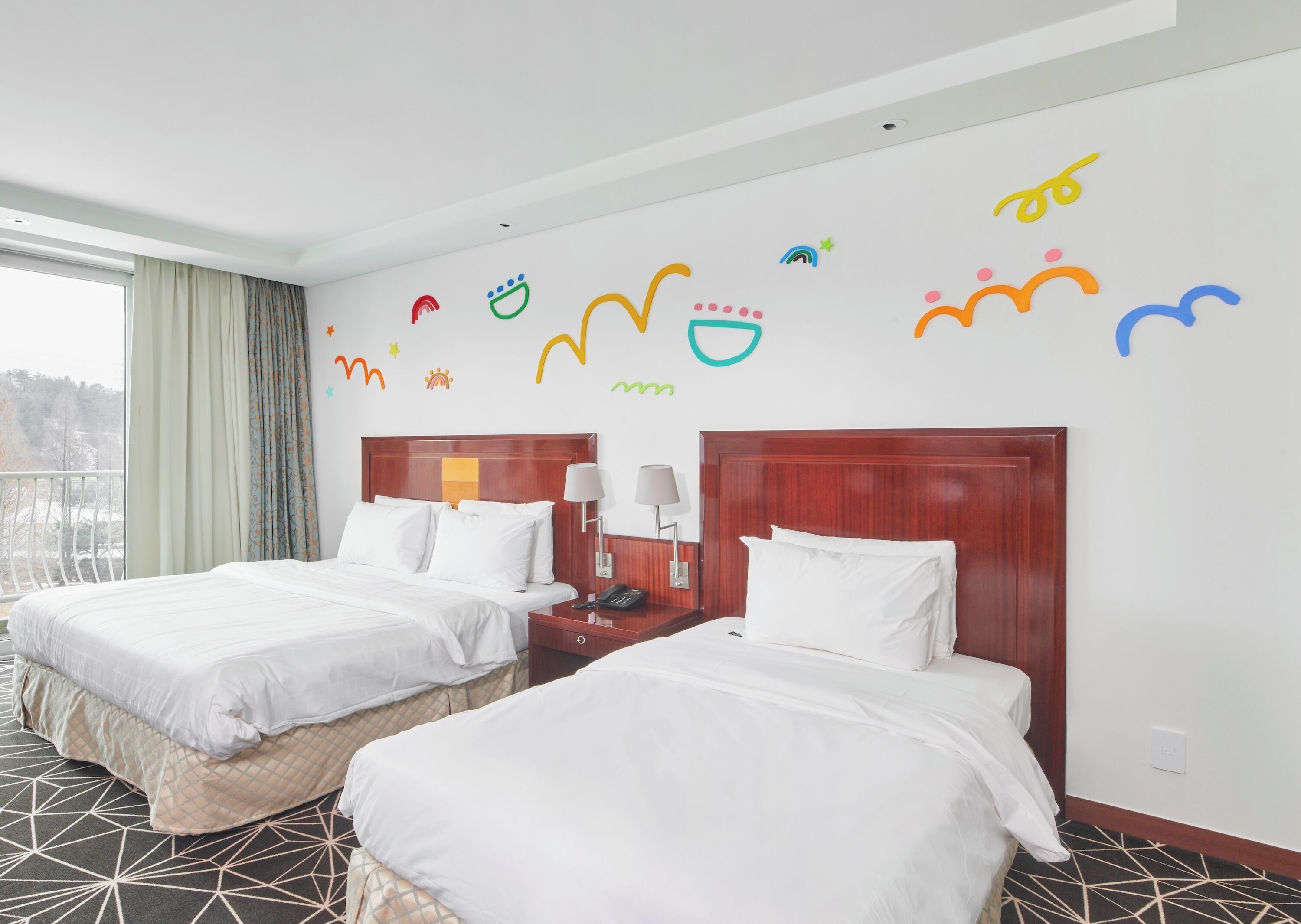 A hotel room with two beds, a wooden headboard, a nightstand with a lamp, and a window with curtains.
