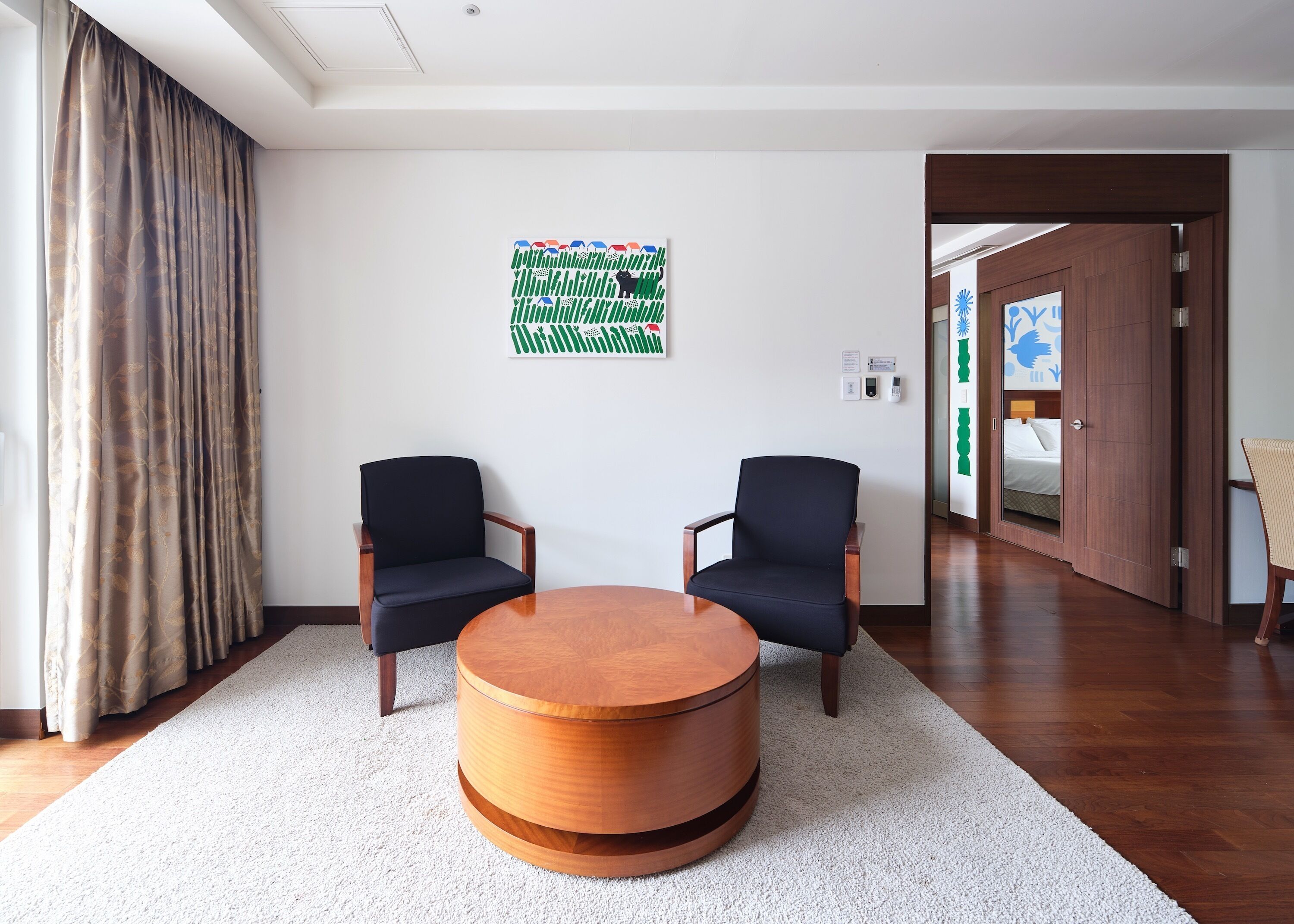 A modern hotel room with two dark blue armchairs, a wooden coffee table, and a large abstract painting on the wall.