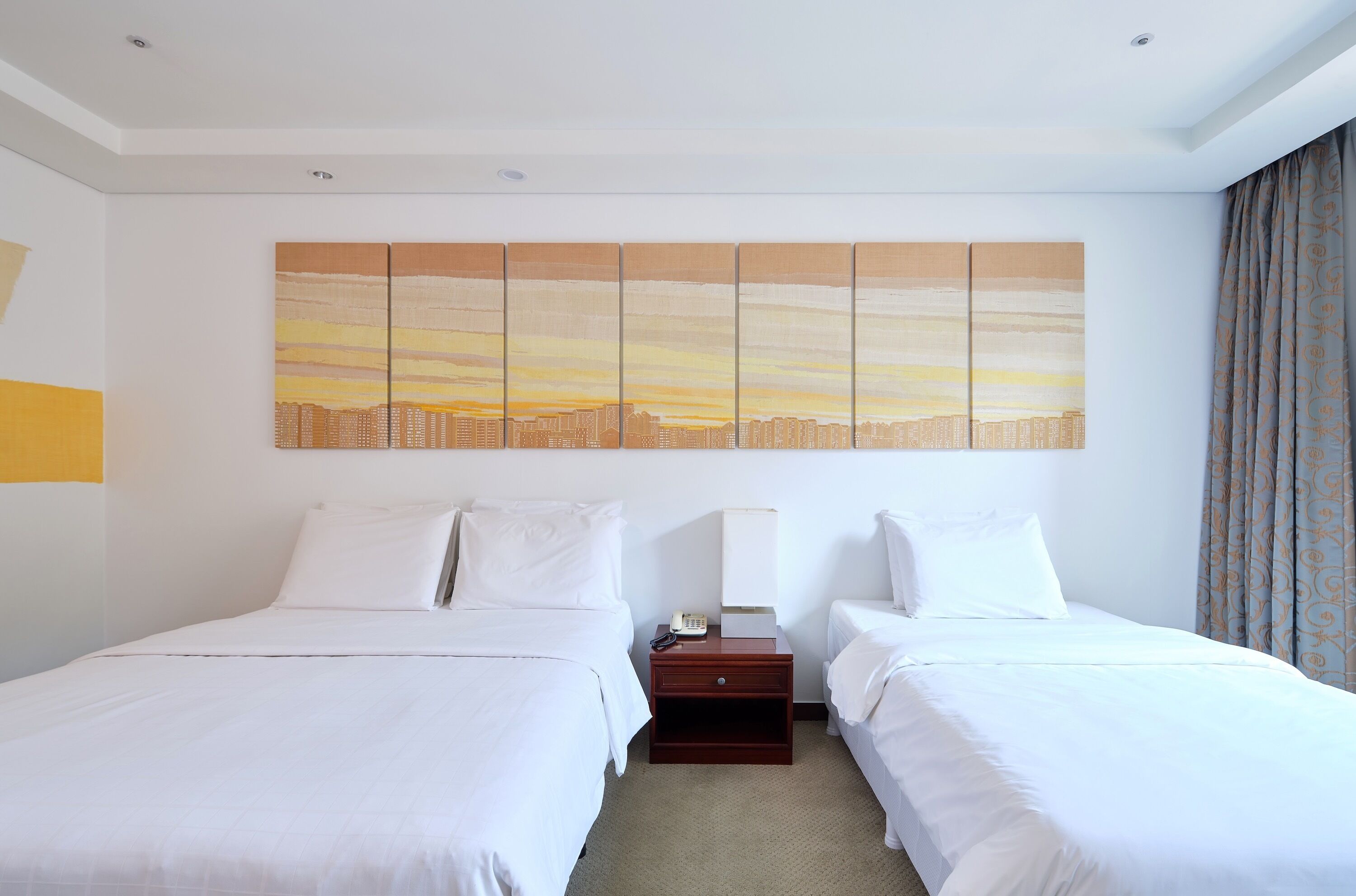 Habitación de hotel con dos camas, una mesita de noche con lámpara y un cuadro grande de un paisaje urbano en la pared.