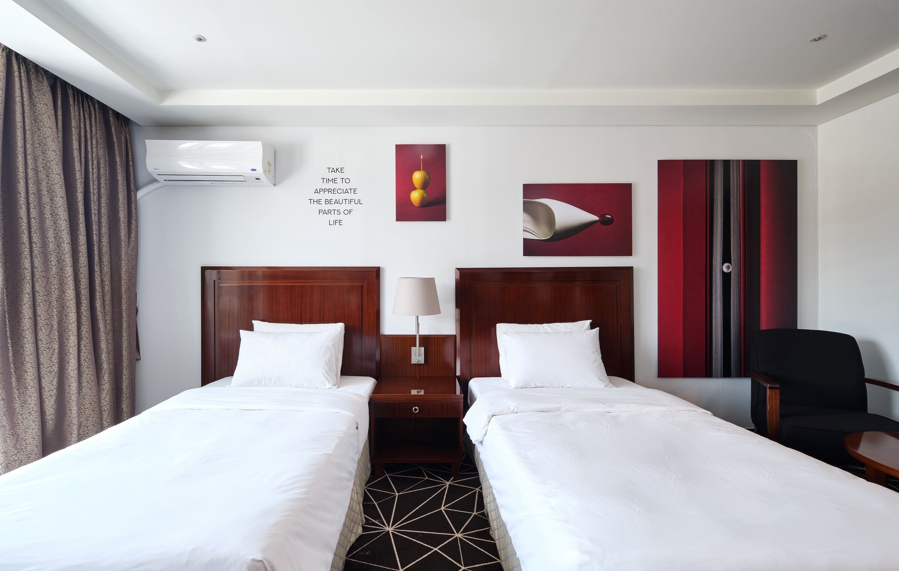A hotel room with two beds, a desk, a chair, a lamp, and artwork on the walls.