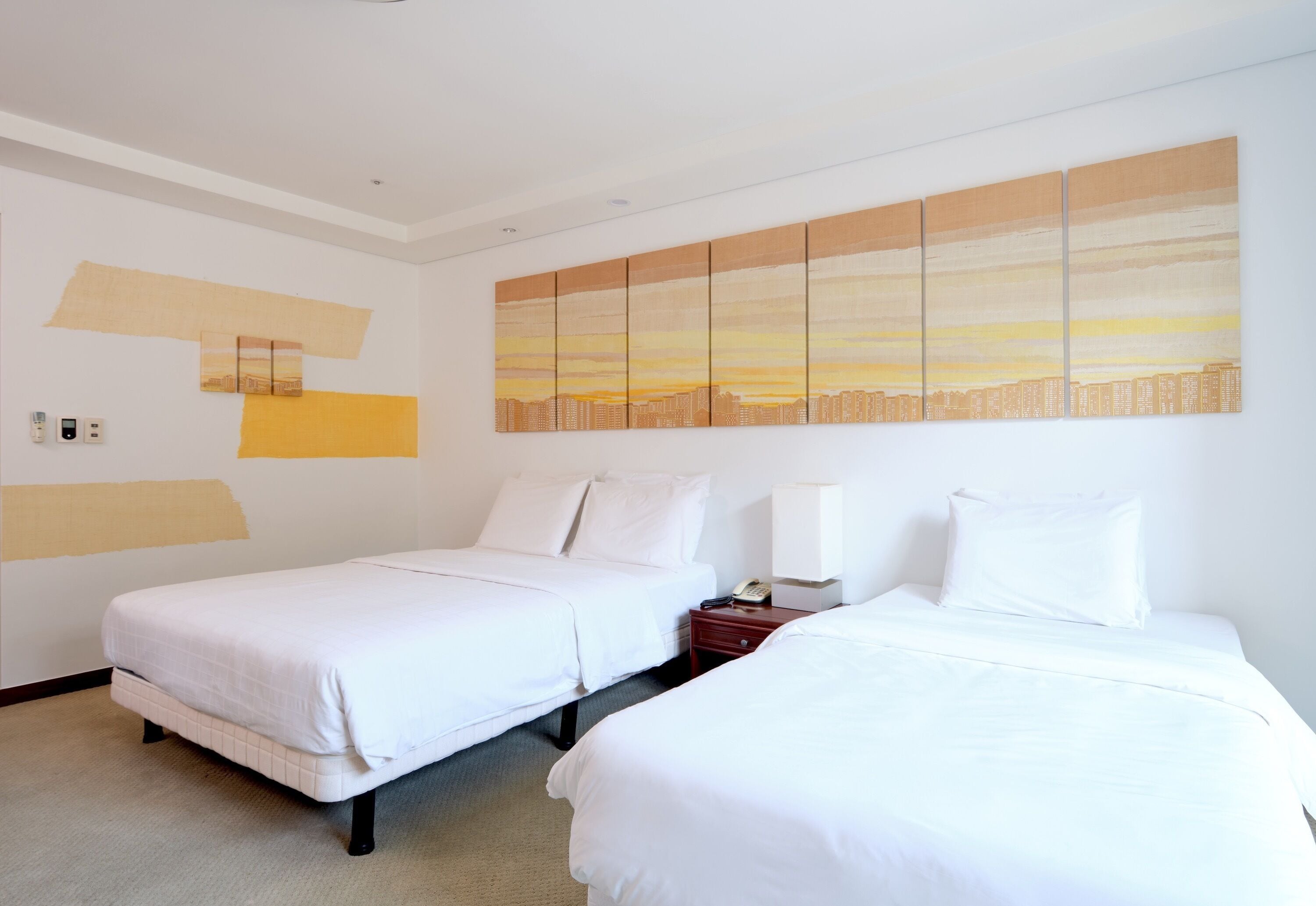 A hotel room with two beds, a cityscape painting, and a nightstand with a phone.