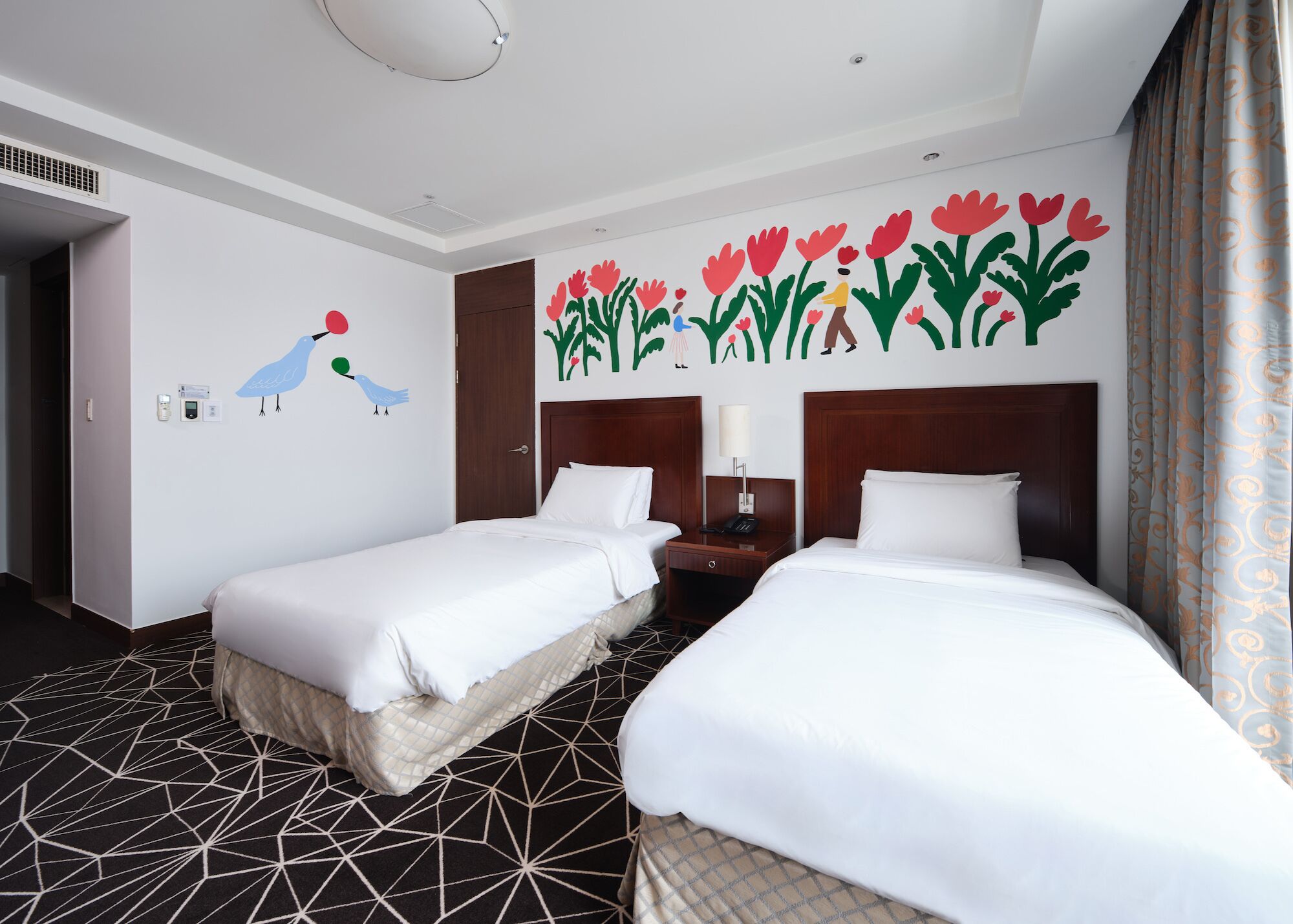 A hotel room with two beds, a nightstand, a wall with a floral mural, and a geometric patterned floor.